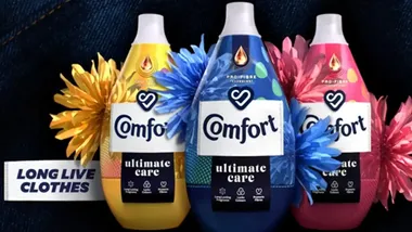 Bottles of Comfort fabric conditioner in yellow, blue and pink lined up. The background is dark denim material. Text reads: Long live clothes.